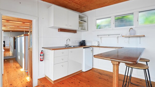 A compact kitchen with white cabinets, a wooden countertop, and a small refrigerator. There is a window with a view of greenery outside, and a fire extinguisher mounted on the wall. - Boatshed Waterfront Huskisson (Huskisson)