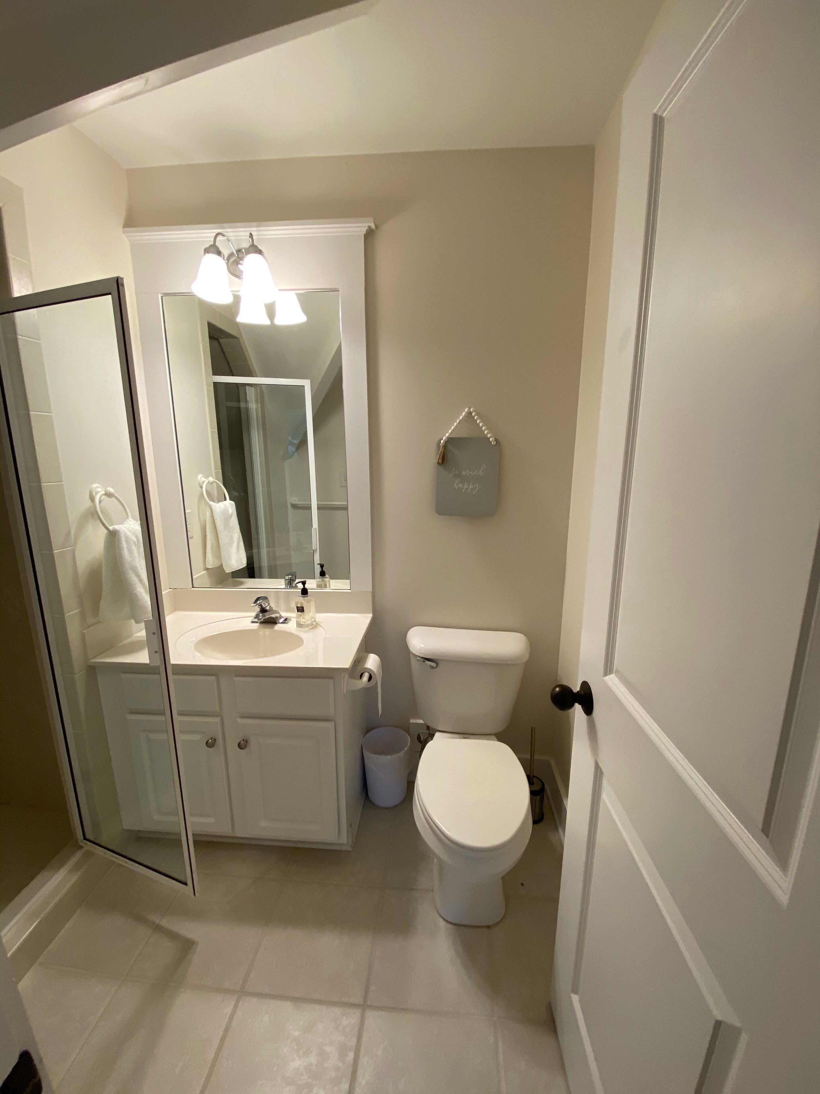 A bathroom with a white sink, mirror, and toilet. There is a towel rack with towels, a trash can, and a small sign on the wall.