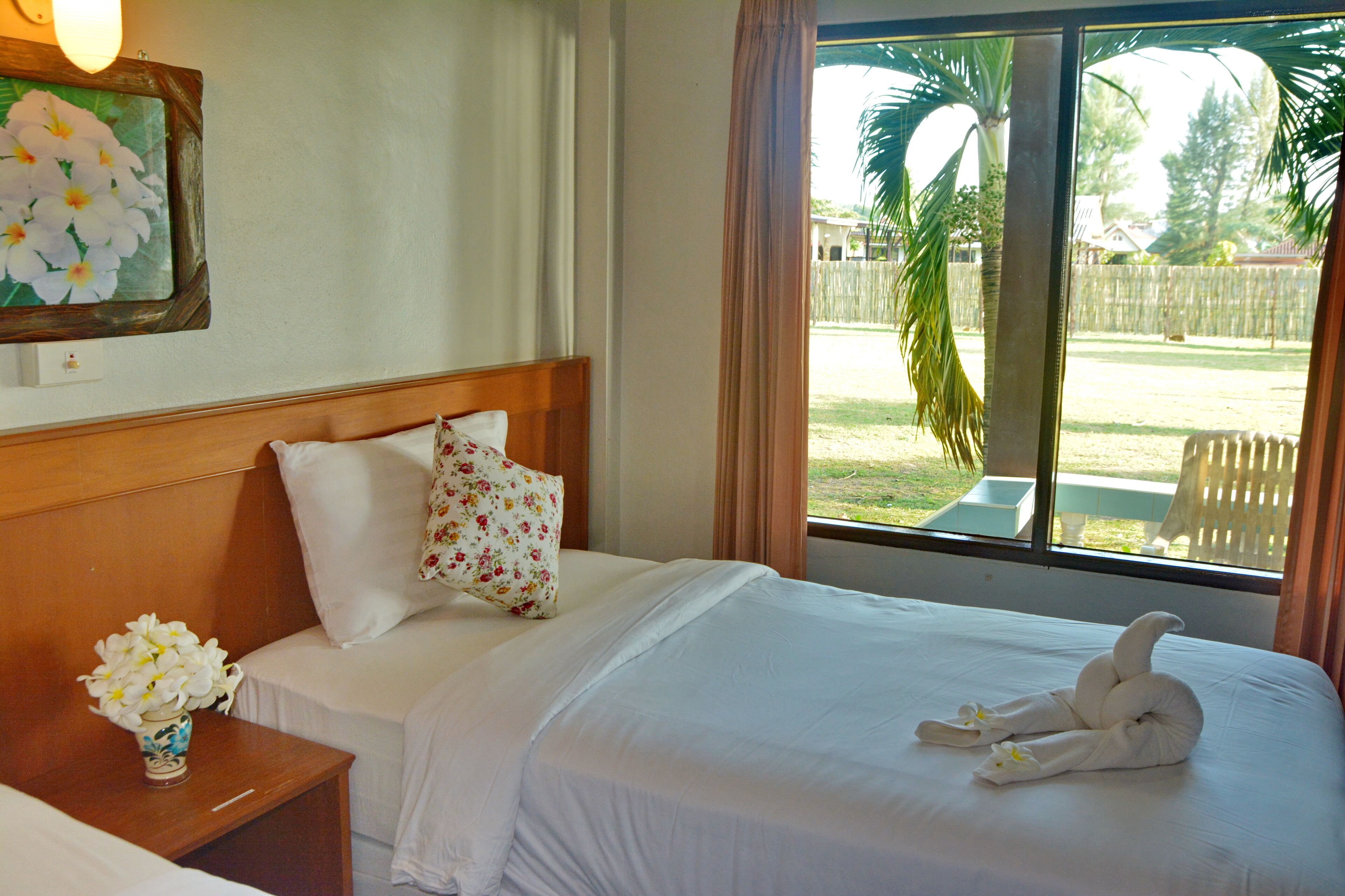 A hotel room with a bed, a nightstand, a vase with flowers, and a window with a view of the outdoors.
