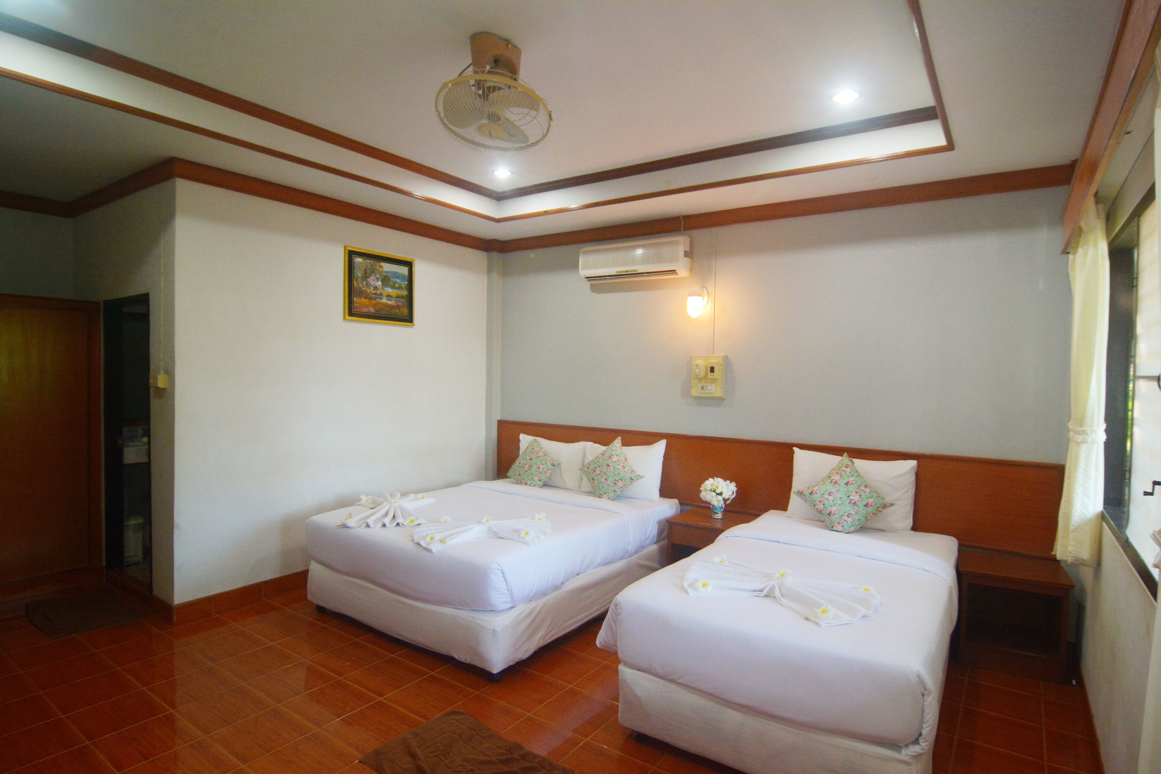 A hotel room with two beds, a ceiling fan, an air conditioning unit, and a small bedside table with a vase of flowers.