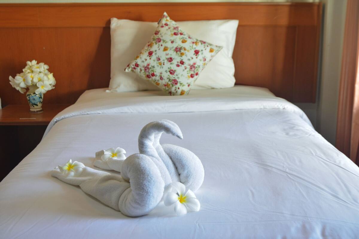 Ein ordentlich bezogenes Bett mit einem blumigen Kopfkissen, einer Vase mit Blumen und einem zu einem Schwan geformten Handtuch.