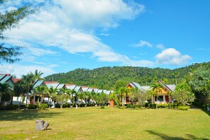 A row of bungalow-style accommodations with sloped roofs, surrounded by well-maintained lawns and tropical vegetation, set against a backdrop of a lush green hill.