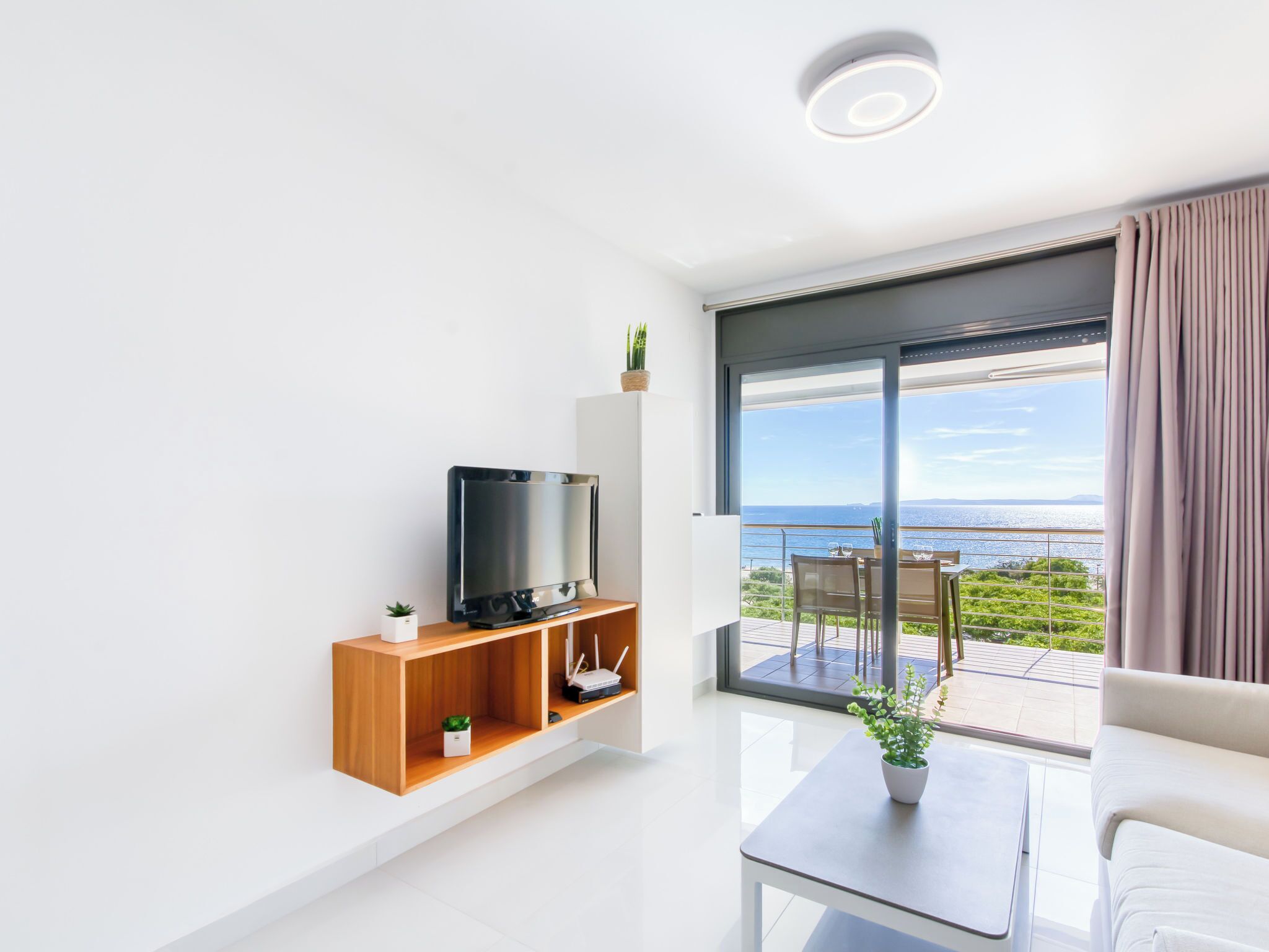 A modern living room with a television on a wooden shelf, a small table with a plant, and a view of the sea through large sliding doors.