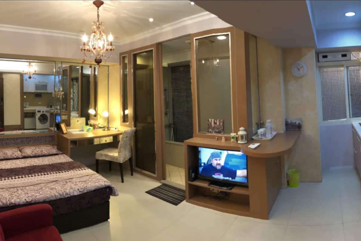 Quarto de hotel com uma cama, uma escrivaninha com cadeira, televisão e cozinha compacta.