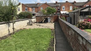 Un quartier résidentiel composé de maisons mitoyennes, d’un étroit sentier et d’un petit jardin agrémenté d’un banc et d’un arbre.