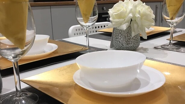 Une table à manger dressée avec des bols blancs et des serviettes jaunes, un vase rempli de fleurs blanches, et une cuisine moderne en arrière-plan.