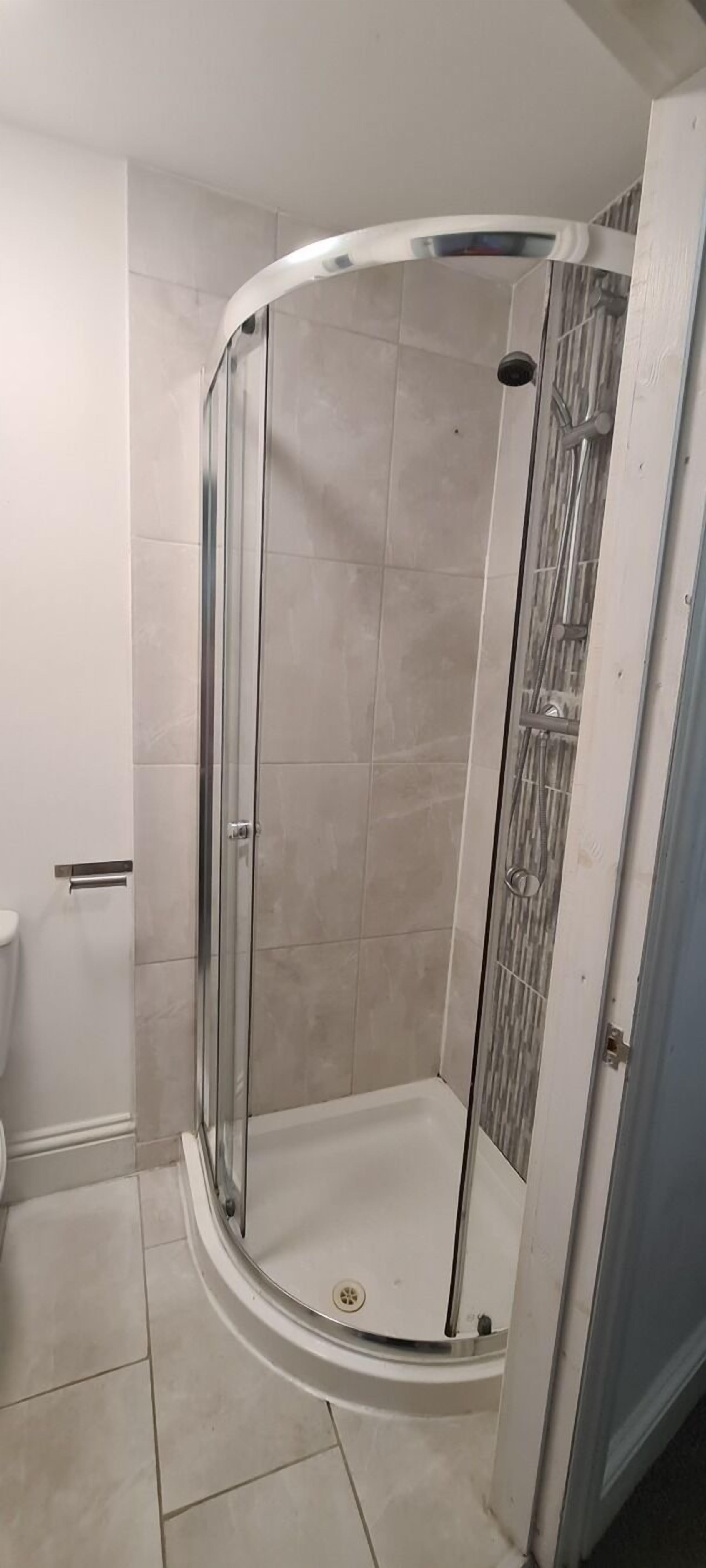 A glass-enclosed shower stall with a white tiled floor and a door leading to another room.