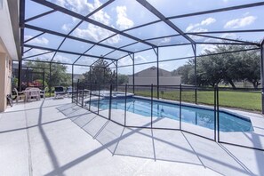 A covered outdoor swimming pool with a metal frame and a clear roof, surrounded by a concrete deck and a patio area with outdoor furniture. - Windsor_palms_8144 (Kissimmee)