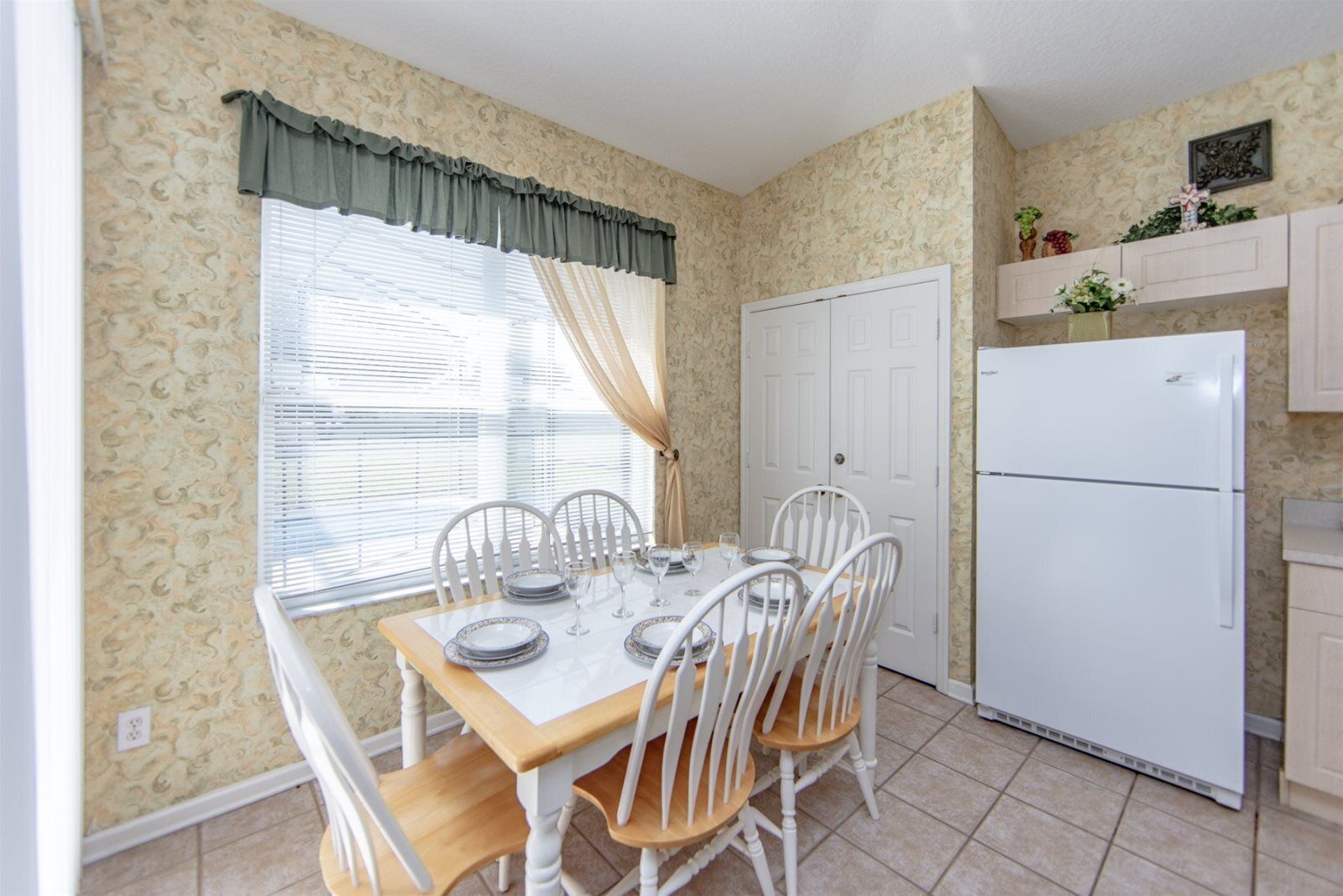 A dining area with a table set for four, white chairs, a white refrigerator, and a window with curtains.
