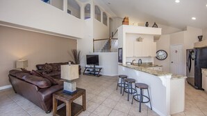 A spacious living room with a brown leather sofa, a television, and a small coffee table. There is a kitchen area with a bar and stools, a refrigerator, and a clock on the wall. - Windsor_palms_8169n (Kissimmee)