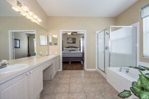 Une salle de bain spacieuse avec un grand meuble-lavabo, une douche à l’italienne et une chambre séparée, visible à travers une porte ouverte.