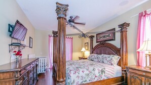 A four-poster bed with floral bedding, a wooden dresser with drawers, a nightstand with a lamp, and a ceiling fan. - Emerald_island_2703 (Kissimmee)