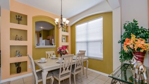 A dining area with a white table set for four, surrounded by beige chairs. There is a chandelier above the table, and a potted plant with orange flowers to the right. - Emerald_island_2703 (Kissimmee)