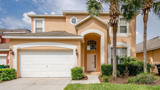 A two-story house with a white garage door, a front entrance with a small porch, and a palm tree in the front yard. - Emerald_island_2703 (Kissimmee)