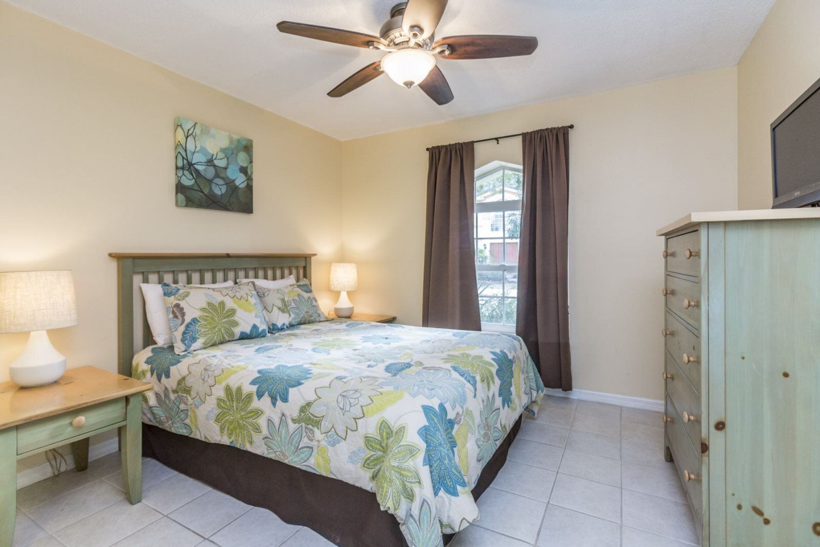 A bedroom with a bed, a nightstand, a dresser, a ceiling fan, and a window with curtains.