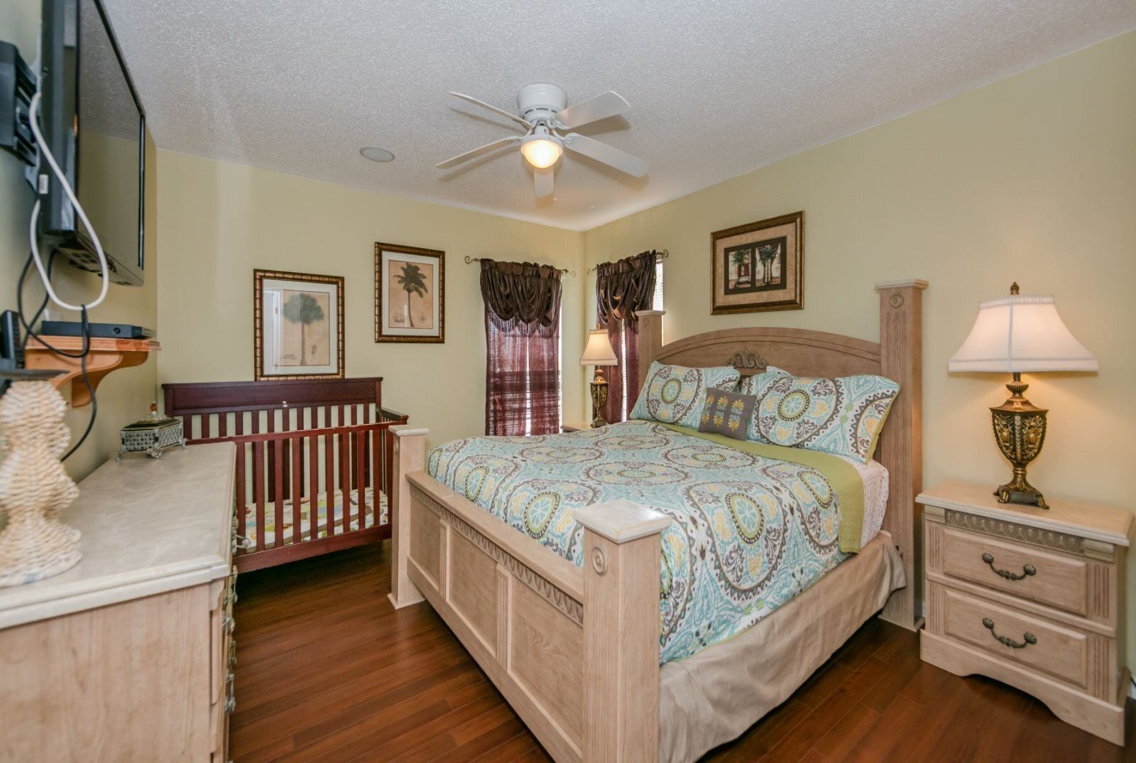 A bedroom with a wooden bed, a crib, a nightstand with a lamp, and a ceiling fan.