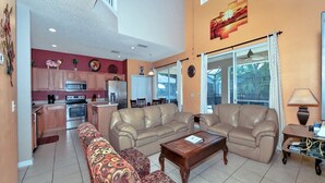 A spacious living room with a beige sofa set, a patterned armchair, and a coffee table. A kitchen with modern appliances is visible in the background. - Emerald_island_8520 (Kissimmee)