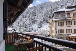 A balcony with a table and chairs, overlooking a snow-covered mountain and buildings. - Le Bellevue 08 Cozy Alpine Apartment | Pool & Mountain Views, Argentière (Chamonix), France (Argentière (Chamonix))