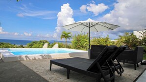 Zona de piscina con sillones de descanso y una sombrilla, con vistas a un cuerpo de agua y montañas.