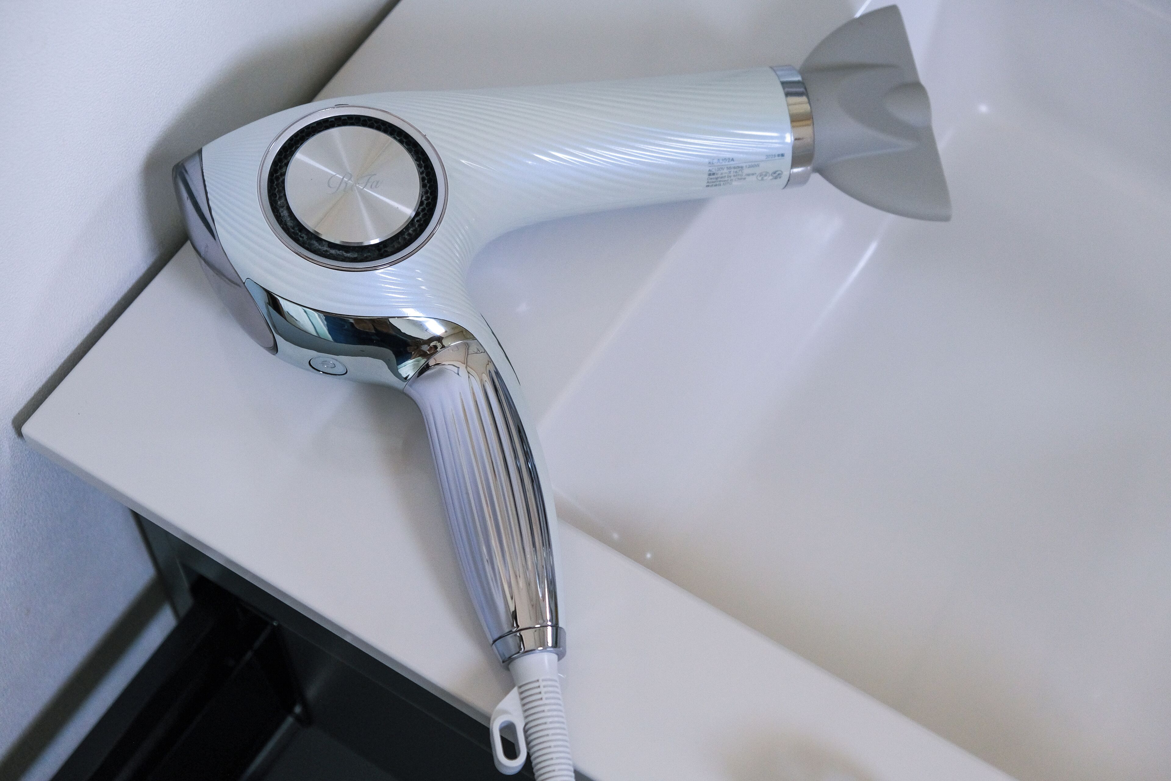 Un sèche-cheveux doté d’un manche métallique et d’un corps blanc, posé sur une surface blanche.