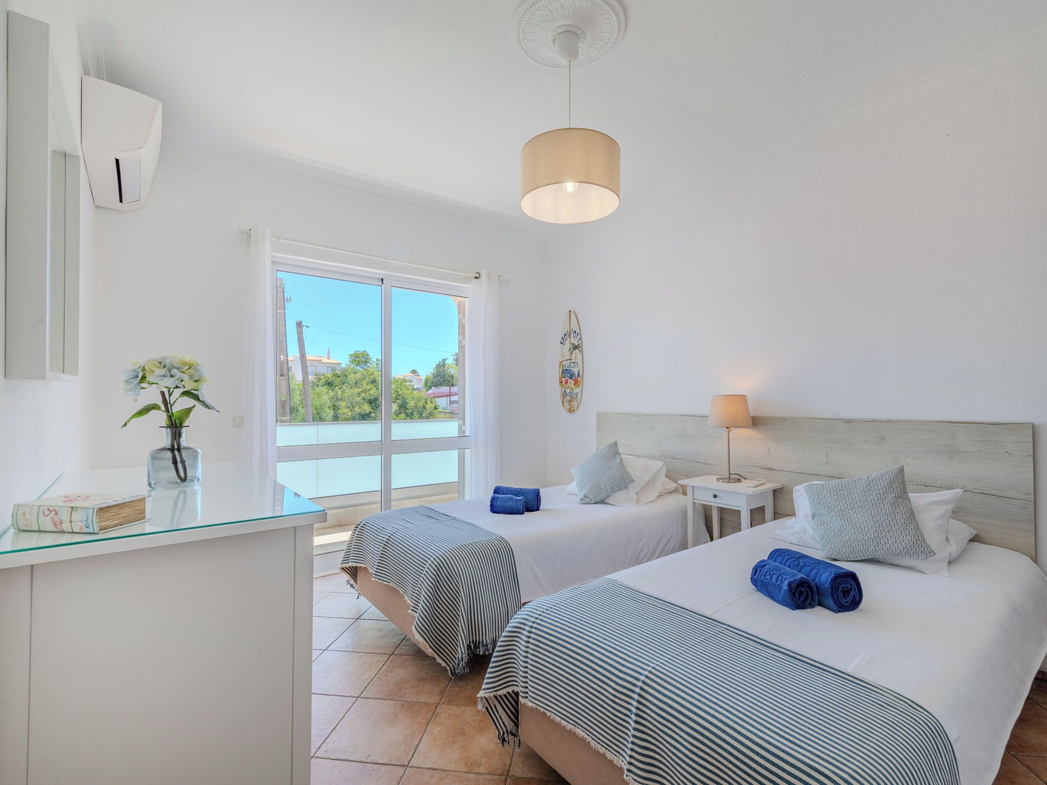 A hotel room with two beds, a glass desk, a vase with flowers, a ceiling fan, and a view of the outside.