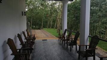 Uma área de estar coberta ao ar livre, com fileiras de cadeiras de madeira, piso pavimentado e vista para uma área gramada com árvores.