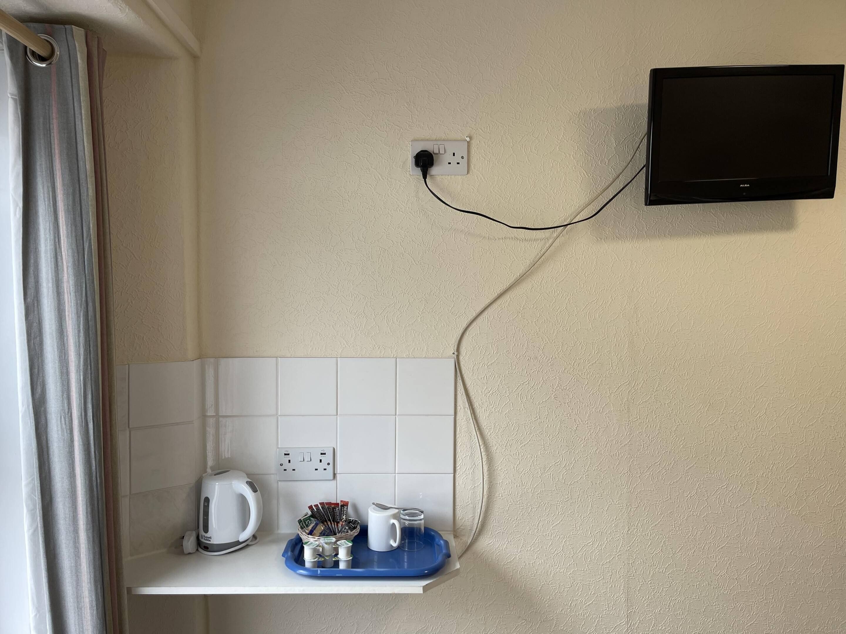 A bathroom with a shower curtain, a white tiled wall, a mounted TV, and a shelf with a kettle, a mug, and toiletries.