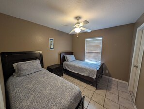 Un dormitorio con dos camas, un ventilador de techo, una ventana con persianas y un cuadro en la pared.