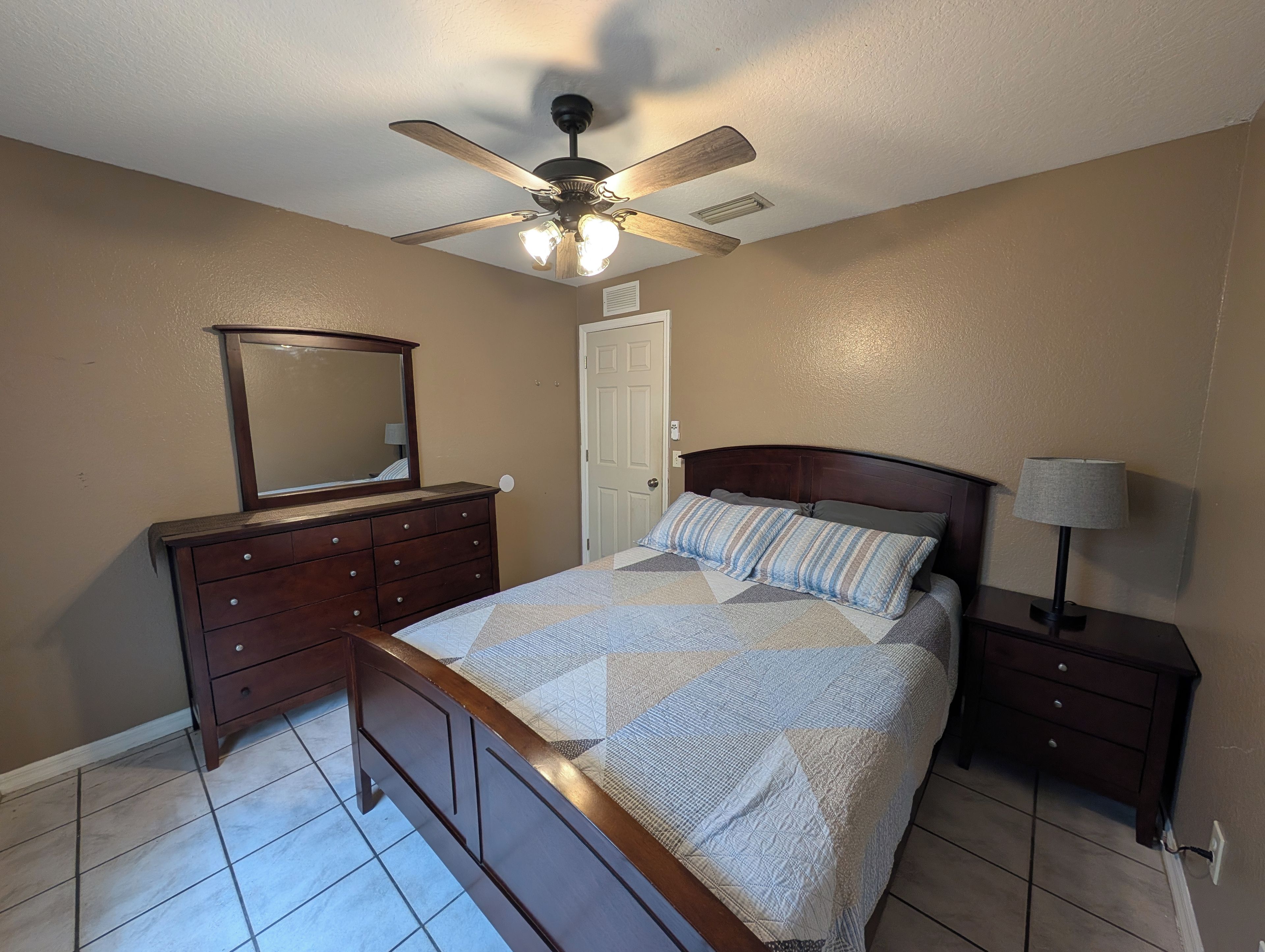 Un dormitorio con cama, cómoda, espejo, ventilador de techo y lámpara de noche.