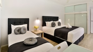 A hotel room with two beds, each with a black headboard, white bedding, and decorative pillows. There is a small table with a chair, and a window with frosted glass panels. - Whitsunday Apartment on Level 6 (Whitsundays)