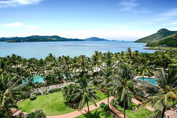 A resort with multiple swimming pools surrounded by palm trees and a view of the sea and mountains.