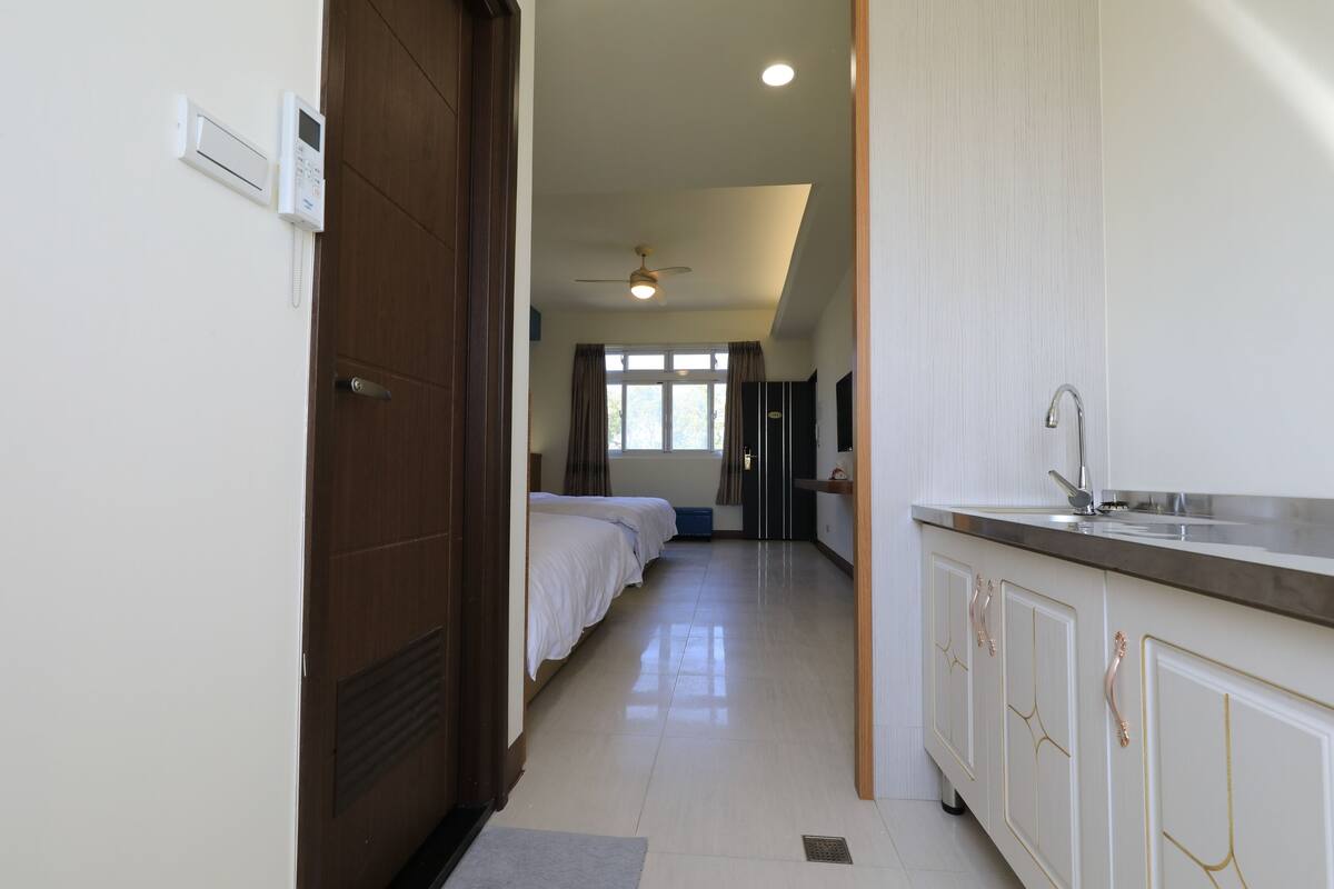 A hotel room with a kitchenette, a bed, a ceiling fan, and a window with curtains.