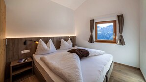 Ein Schlafzimmer mit Bett, Nachttisch, Lampe, Fenster mit Vorhängen und Bergblick.