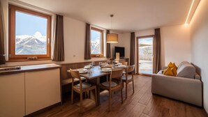 Ein moderner Essbereich mit einem Holztisch, gedeckt für sechs Personen, großen Fenstern mit Bergblick, einer Couch mit Kissen und einem Flachbildfernseher.