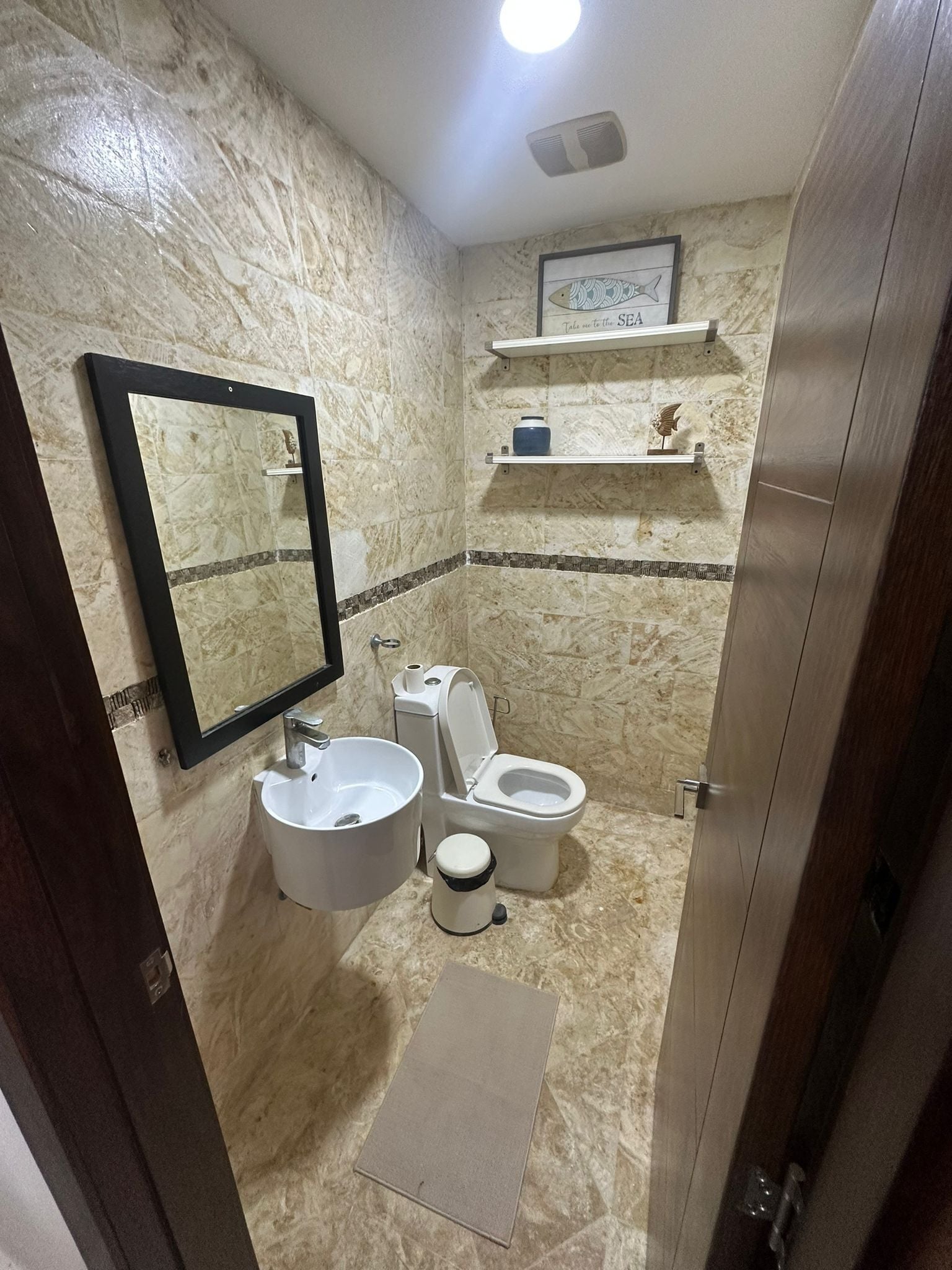 Une salle de bain avec un mur en marbre, un lavabo, des toilettes, un miroir et une étagère contenant des articles de toilette.