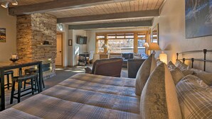 Una habitación acogedora con chimenea de piedra, una cama grande con ropa de cama escocesa, techo de madera y vistas al exterior a través de amplias ventanas.