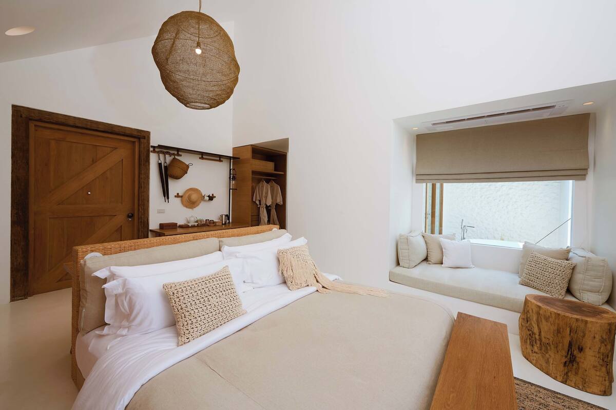 Un dormitorio con una cama grande, un armario de madera, una lámpara colgante, una ventana con vistas y un banco empotrado con cojines.