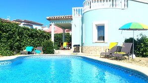 Une piscine entourée de chaises longues et de parasols, le tout bordé de verdure et adossé à un bâtiment d’un bleu clair.