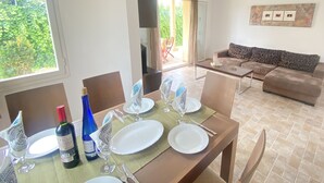 On aperçoit une salle à manger avec une table dressée pour quatre personnes, comprenant des assiettes, des couverts et des verres à vin. Un canapé et une table basse sont visibles au fond.
