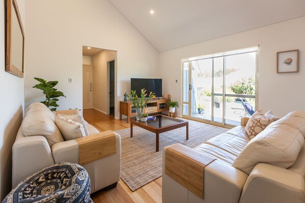 A spacious living room with a high ceiling, a large sliding glass door leading to a balcony, and a flat-screen TV on a wooden stand.