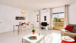 A modern living room with a dining area, a flat-screen TV, and a balcony with curtains.