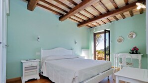 A bedroom with a wooden ceiling, a white bed, a bedside table, a chair, and a view of the outdoors.