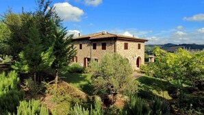 A stone building with a tiled roof, surrounded by greenery and hills in the background.