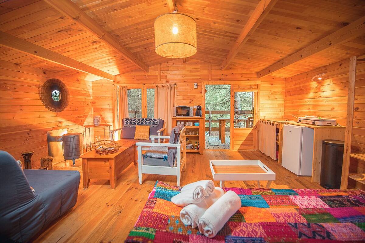 Interior acogedor de una cabaña de madera con sofá, colcha, mesa de comedor y zona de cocina.