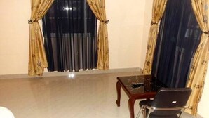 Una habitación con una mesa de madera y una silla negra, dos ventanas con cortinas y un piso de baldosas.