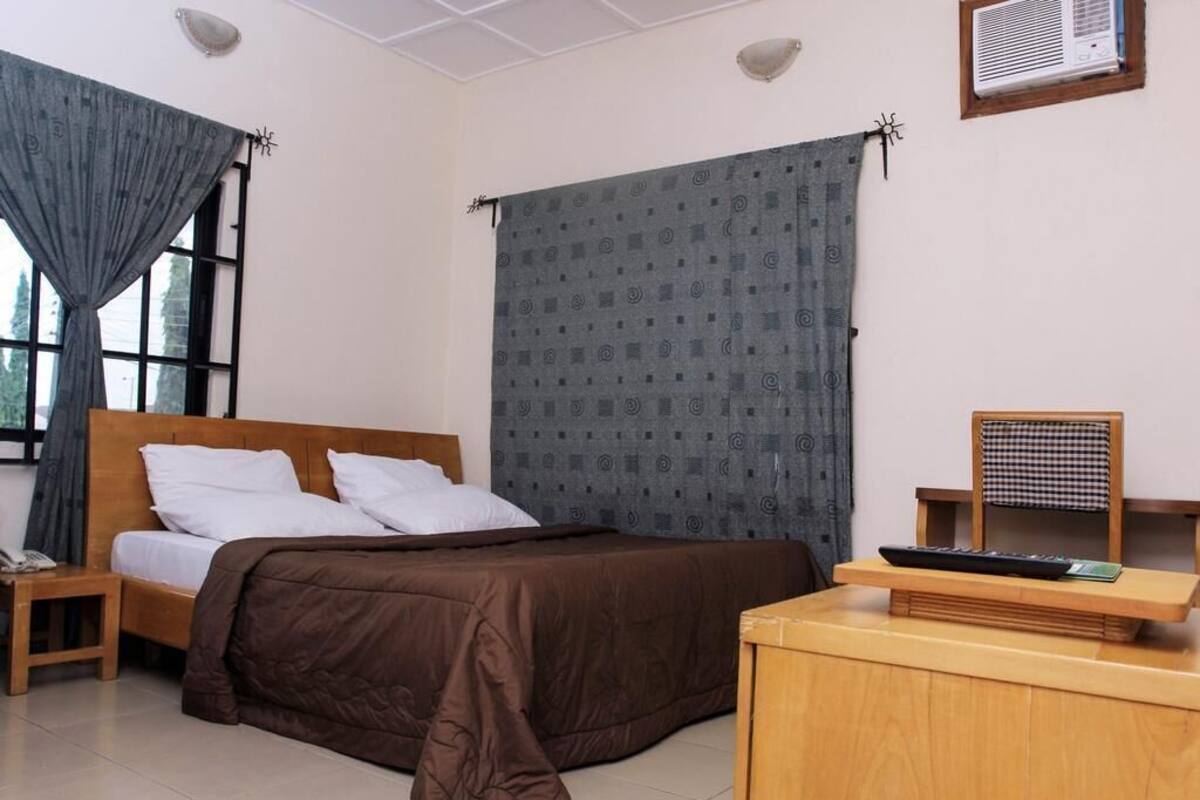 A bedroom with a bed, a desk, a chair, a window with curtains, and a wall-mounted air conditioner.