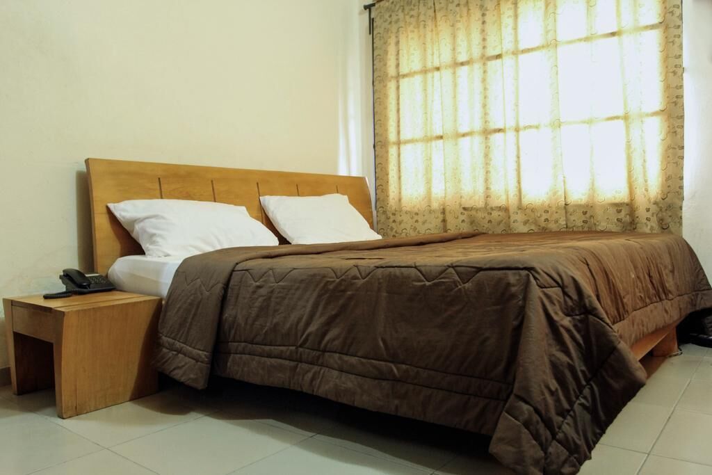 Una cama individual con cabecera de madera, dos almohadas blancas, una colcha marrón y una mesita de noche de madera con un teléfono.