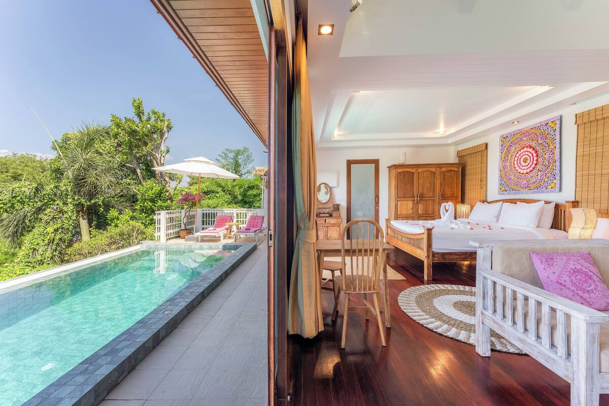 A bedroom with a large bed, a wooden chair, a dining table, and a view of a swimming pool.