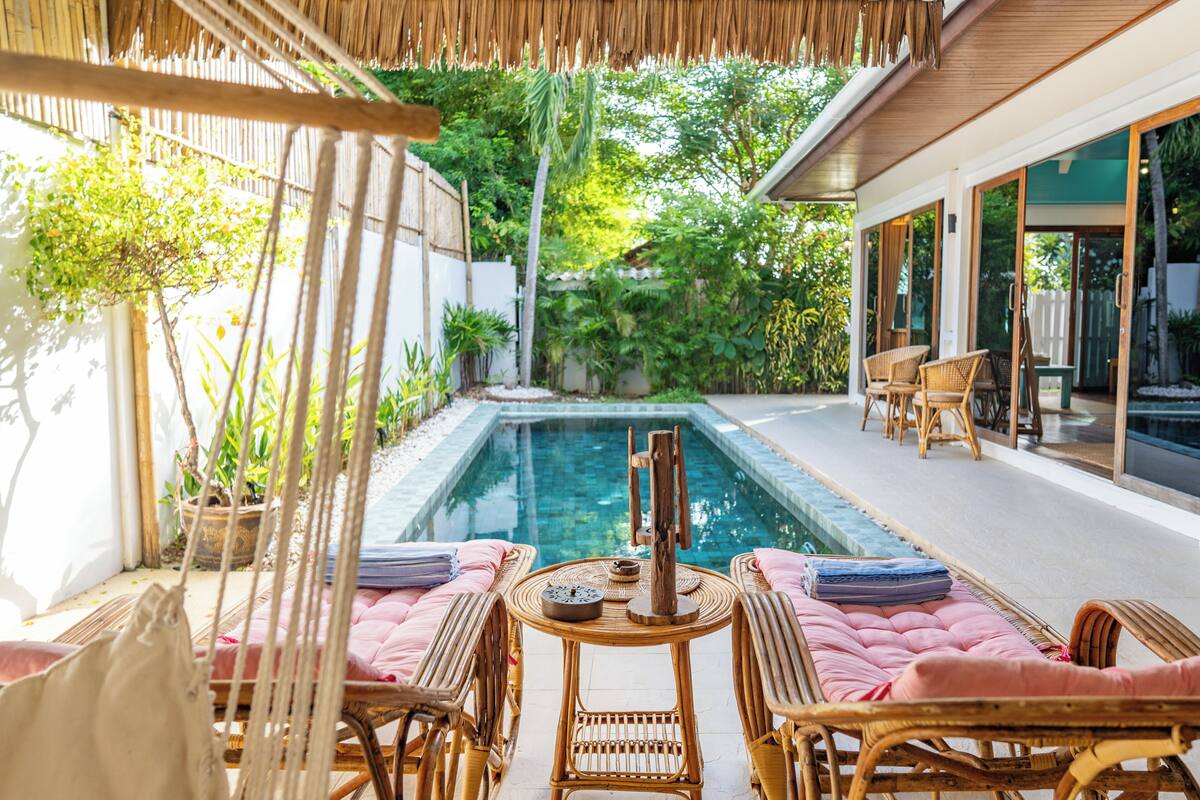 A pool area with wicker furniture, a table with a clock, and a view of the surrounding greenery.