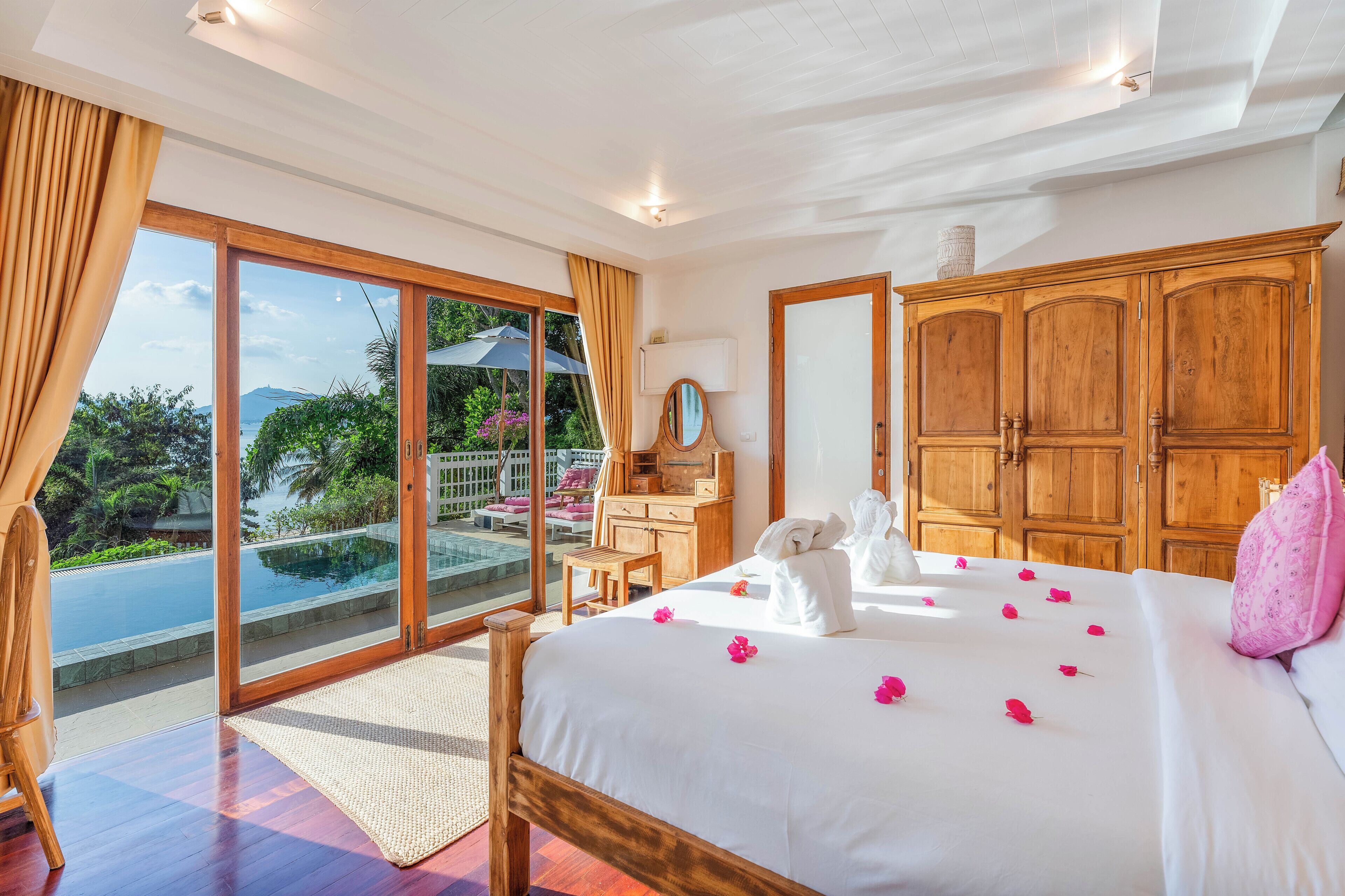 A bedroom with a large bed, wooden wardrobe, and a view of an infinity pool.